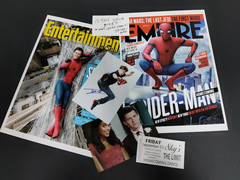May include: A red and blue Spider-Man costume with a red mask. The costume is on a magazine cover with the title "Spider-Man Homecoming". The magazine is on a black surface with a signed photo of Spider-Man and a ticket for a homecoming dance.