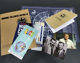 Könnte beinhalten: Ein brauner Ordner mit dem Text "S.H.I.E.L.D." und einem schwarzen Logo. Der Ordner ist geöffnet und es liegen mehrere andere Gegenstände darauf, darunter ein blaues und weißes Ticket, ein brauner Ordner mit dem Text "TOP SECRET", ein blauer und weißer Bauplan und ein Foto von zwei Männern.