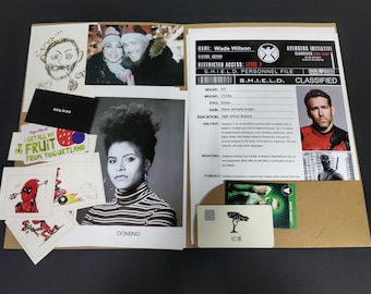 Könnte beinhalten: Ein brauner Ordner mit einem Charakterprofil für Wade Wilson, auch bekannt als Deadpool, von der Avengers Initiative. Das Profil enthält Informationen über seine Größe, sein Gewicht, seine Augen, seine Haare, seine Ausbildung, seine Fähigkeiten, seine Waffen und seine Kräfte. Der Ordner enthält auch ein Foto von Deadpool in seinem rot-schwarzen Kostüm.