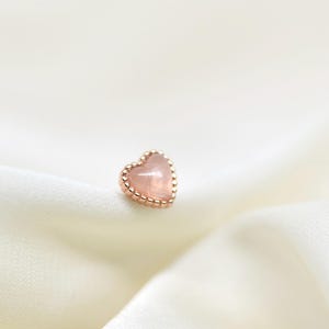 Op de afbeelding: Een hartvormige oorbel met een roségouden zetting en een lichtroze edelsteen. De oorbel is geplaatst tegen een achtergrond van zachte, witte stof, wat een delicate en vrouwelijke esthetiek creëert.