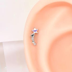 May include: A silver ear piercing featuring a star design with iridescent accents and a dangling chain of smaller stars. The earring is shown on a model ear, showcasing its placement and style. The jewelry is set against a soft, neutral background.
