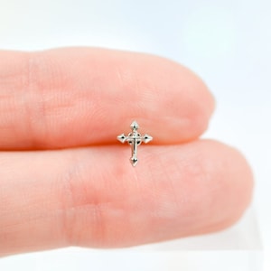 May include: A small, silver-toned metal stud earring shaped like a cross. The cross features a central vertical bar intersected by a horizontal bar, with pointed, decorative ends on each of the four arms, creating a detailed, symmetrical design.