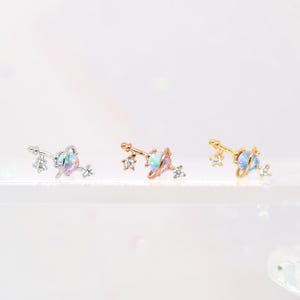 May include: Three pairs of celestial-themed stud earrings. Each earring features a planet design with a central iridescent stone and small star accents. The earrings are in silver, rose gold, and gold tones.