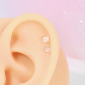 May include: A close-up of a fake ear with two clear stud earrings in the helix piercing.