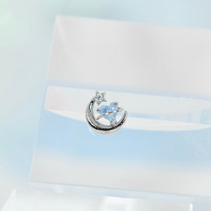 May include: A silver-toned ring featuring a delicate crescent moon and a small star, centered with a light blue heart-shaped stone. The design is polished and metallic, showcasing a celestial motif with a clear, pale blue gemstone accent.