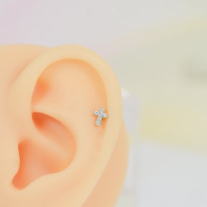 May include: A close-up of a fake ear with a small, diamond-encrusted cross earring. The earring is made of gold and has a simple, elegant design.