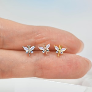 May include: Three dragonfly-shaped earrings with iridescent white wings and clear crystal accents. The earrings are shown on a finger, with the silver, rose gold, and gold versions of the earring.