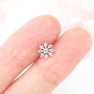 Puede incluir: Un pendiente de botón de plata en forma de copo de nieve con piedras de circonita cúbica transparentes.