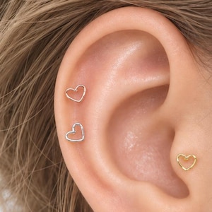 May include: Three small heart-shaped stud earrings worn in the ear. Two earrings are positioned on the upper cartilage, one in rose gold and one in silver. A third gold heart-shaped earring is placed in the conch area of the ear.