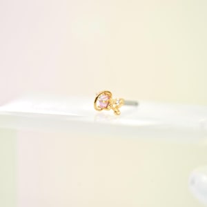 May include: A gold-colored nose stud featuring a pink gemstone and small, clear accent stones. The stud is set on a silver-colored post. The jewelry is displayed on a white surface.