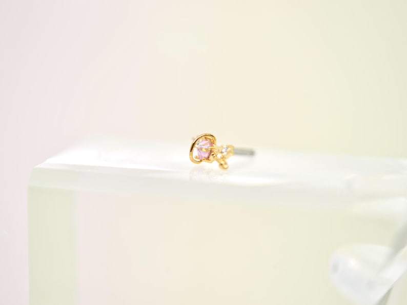 May include: A gold-colored nose stud featuring a pink gemstone and small, clear accent stones. The stud is set on a silver-colored post. The jewelry is displayed on a white surface.