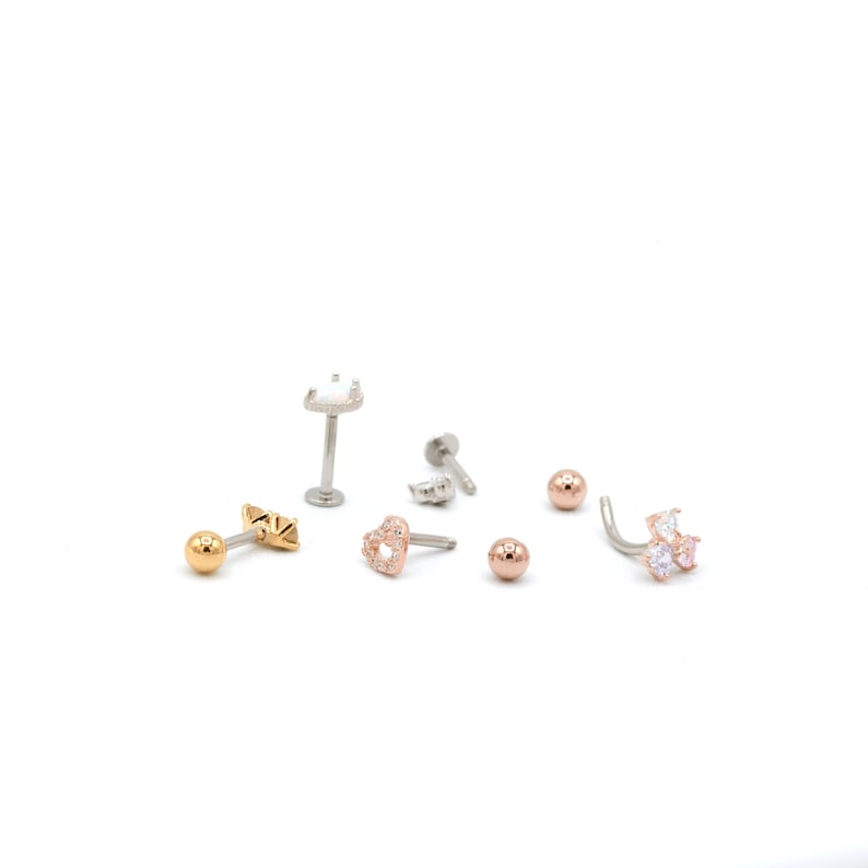 May include: A collection of body jewelry, including gold and silver-toned earrings. The earrings feature various designs, such as a heart, a ball, and a cluster of gemstones. The jewelry is displayed on a white background.