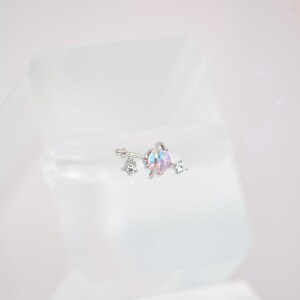 May include: A silver-colored belly button ring with a floral design. The ring features a central iridescent gem with pink and blue hues, flanked by clear, sparkling crystals. The jewelry is set against a white background.