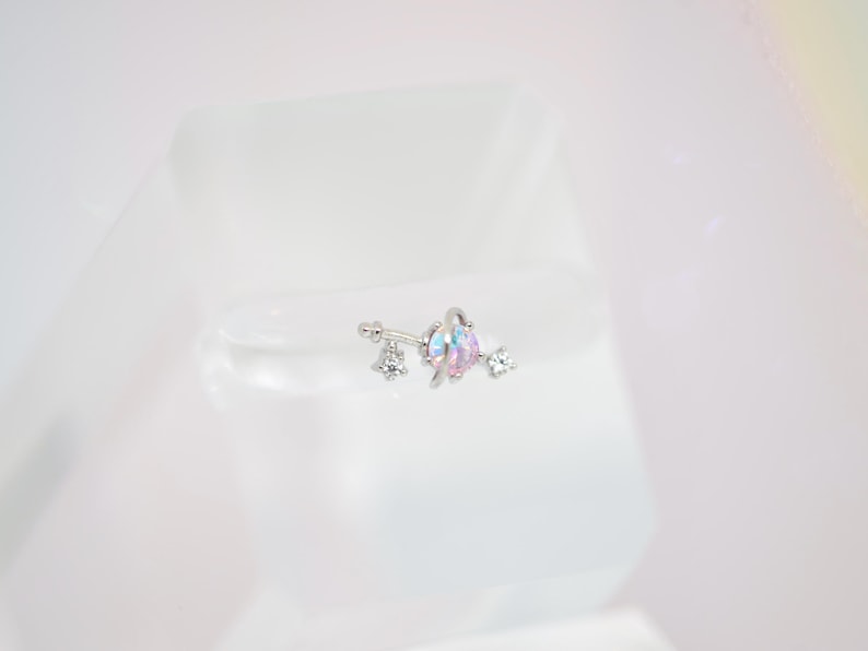 May include: A silver-colored belly button ring with a floral design. The ring features a central iridescent gem with pink and blue hues, flanked by clear, sparkling crystals. The jewelry is set against a white background.