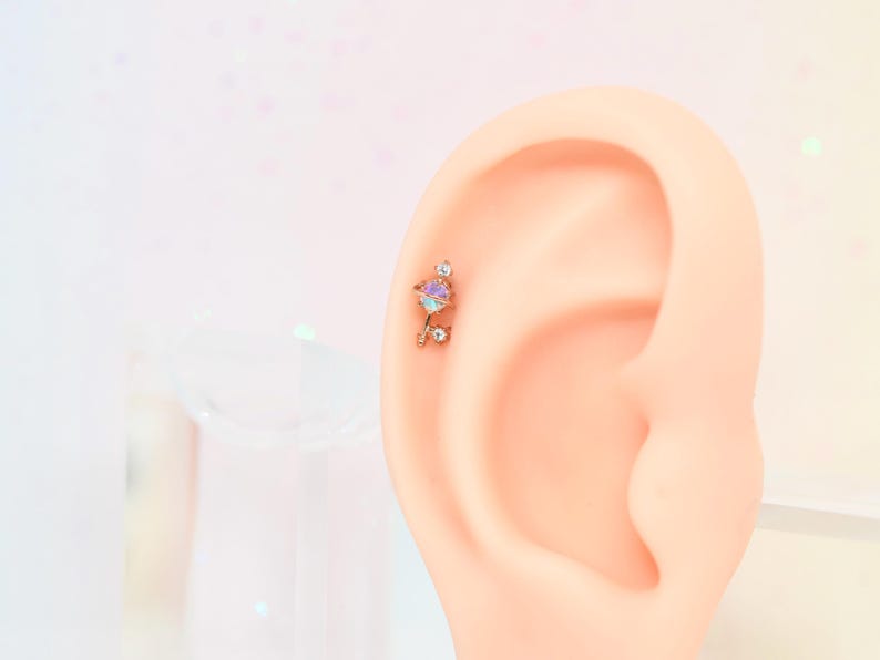 May include: A rose gold helix piercing with a colorful, iridescent gemstone and small clear crystals. The earring is attached to a light-skinned ear, set against a soft, blurred background.