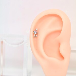 May include: A silver ear piercing with a cluster of iridescent gemstones and small clear crystals. The jewelry is attached to a model ear, showcasing the design and placement of the earring. The background is a soft gradient of pink and white.