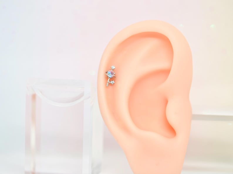 May include: A silver ear piercing with a cluster of iridescent gemstones and small clear crystals. The jewelry is attached to a model ear, showcasing the design and placement of the earring. The background is a soft gradient of pink and white.