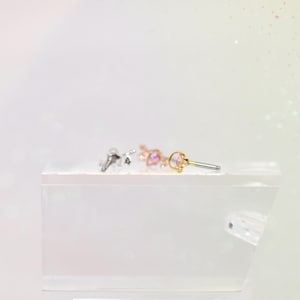 May include: Three small, decorative earrings with a celestial theme are displayed on a white surface. The earrings are silver, rose gold, and gold, each featuring a small, colorful gemstone. The earrings are designed for body piercing.