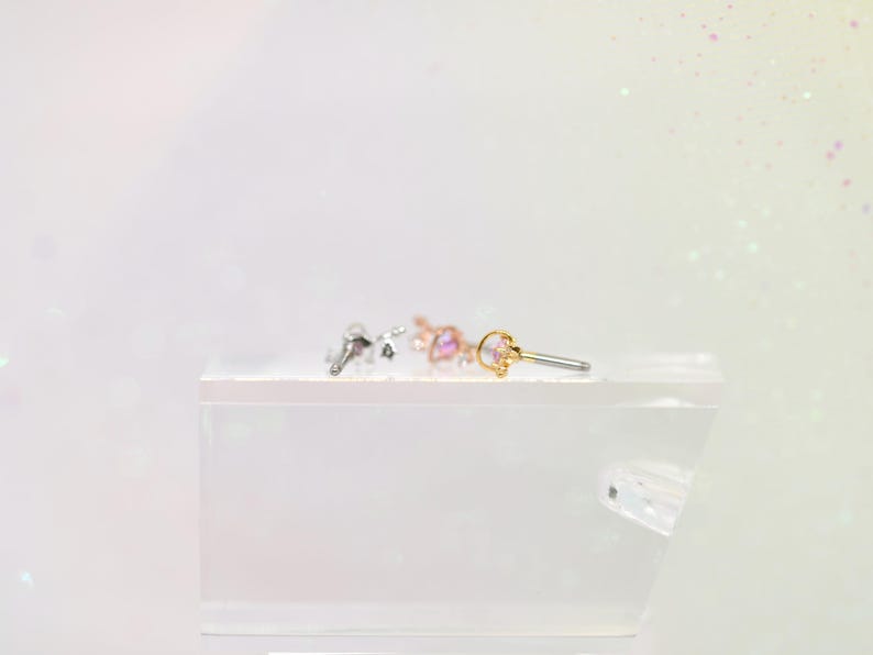 May include: Three small, decorative earrings with a celestial theme are displayed on a white surface. The earrings are silver, rose gold, and gold, each featuring a small, colorful gemstone. The earrings are designed for body piercing.