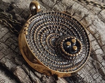 May include: A circular pendant necklace with a bronze finish. The pendant features a concentric circle design with a central black dot surrounded by three smaller black dots.