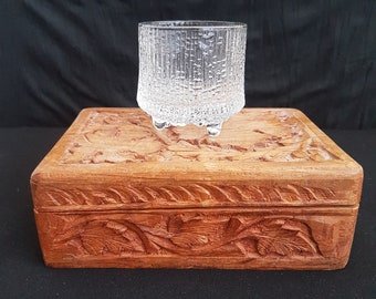 May include: A clear glass with a textured surface and three legs sits on top of a wooden box with intricate carvings. The box is a rich brown color.