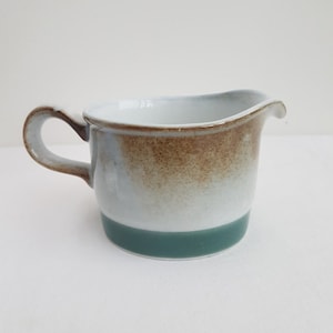 May include: A white ceramic creamer with a brown glaze and a teal band around the top. The creamer has a rounded body and a handle.