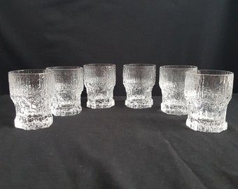 May include: A set of six clear glass tumblers featuring a textured, bark-like exterior pattern. Each glass has a cylindrical shape with a slightly wider rim and a heavy, thick base, showcasing a rugged, organic surface finish throughout the glass.