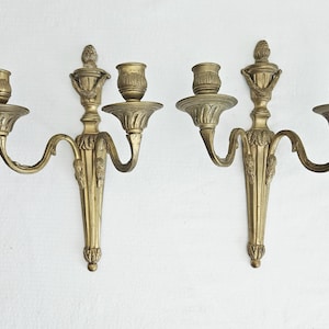 Pode incluir: Um par de arandelas de parede em metal dourado, cada uma com dois suportes para velas com bases estriadas. A placa traseira vertical central é adornada com motivos de folhas e rematada com um topo, enquanto braços curvos sustentam os copos.