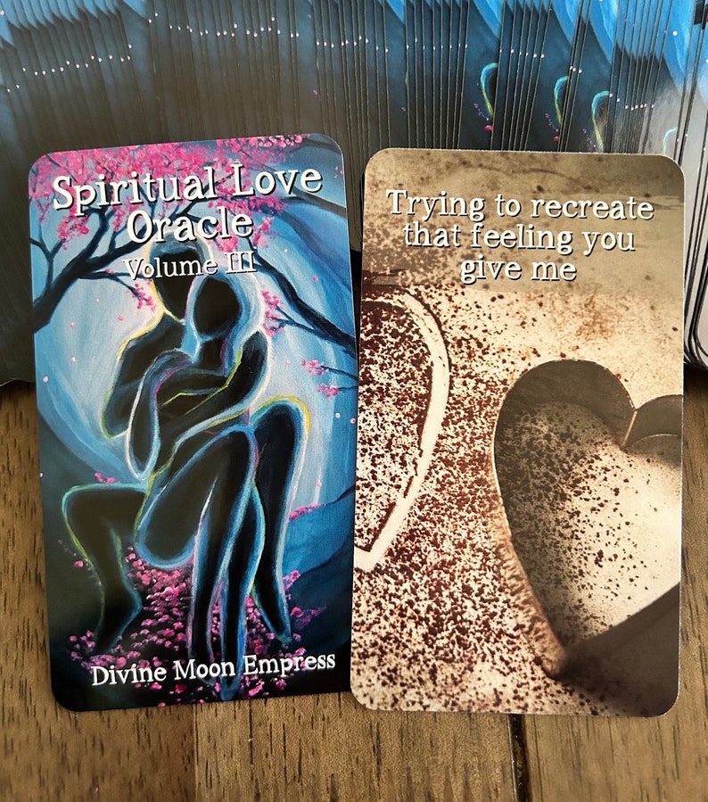 May include: Two oracle cards from the Spiritual Love Oracle deck, Volume III. The first card features a blue and purple illustration of two figures embracing, with pink blossoms in the background. The text on the card reads "Divine Moon Empress". The second card features a brown and white image of a heart shape with the text "Trying to recreate that feeling you give me".