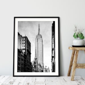 May include: Black and white photograph of the Empire State Building in New York City. The building is seen from a street level perspective, with other buildings lining the street.