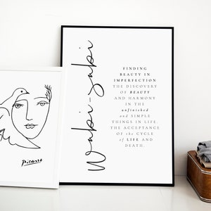 May include: Two black and white framed prints on a white wall. The print on the left is a line drawing of a woman's face with a dove. The print on the right has the text "Waki-jabi" and a quote about finding beauty in imperfection.