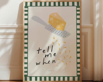 May include: A framed rectangular wall art print featuring a yellow wedge of cheese being grated by a grey metal grater. The design includes the text "tell me when" in black script and a green and white checkered border on a cream background.