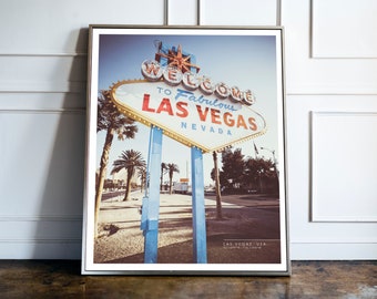 May include: A vintage-style photograph of the iconic "Welcome to Fabulous Las Vegas, Nevada" sign. The sign is lit up with red, white, and blue neon lights against a blue sky. Palm trees and a road are visible in the background.