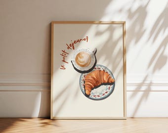 May include: A framed wall art print featuring a top-down illustration of a golden-brown croissant on a floral patterned plate next to a cup of coffee with latte art. The text le petit dejeuner is written in a cursive red font above the items.