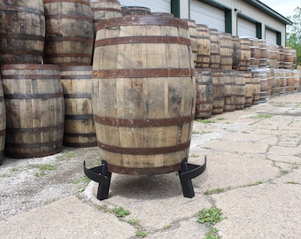 May include: A wooden barrel with metal bands sits on a black metal stand. The barrel is brown and has a weathered look. The stand has three legs and is designed to hold the barrel off the ground.