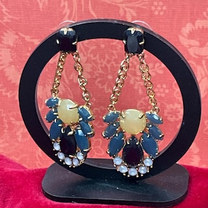 May include: A pair of gold-tone drop earrings featuring a black stud at the top, connected by a gold chain to a cluster of stones. The cluster includes a central yellow stone, surrounded by blue, black, and small white stones in a fan shape.