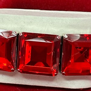May include: A stretch bracelet featuring three large, square-cut, vibrant red faceted stones set in polished silver-tone metal frames. The stones are arranged horizontally, showcasing a deep crimson hue and a reflective, geometric surface design.