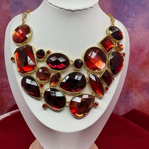 May include: A statement necklace featuring multiple faceted, deep red, translucent stones set in gold-toned metal frames. The stones vary in shape, including round, oval, pear, and rectangular cuts, arranged in a layered, clustered design with a gold chain.