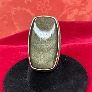 May include: A ring featuring a large, rectangular, polished stone with rounded corners in a muted olive-green and bronze metallic hue. The stone is set within a smooth, polished silver-toned metal bezel that frames the gemstone's surface.