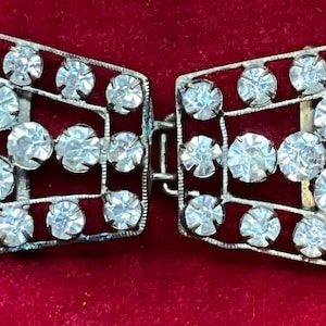 May include: A silver-toned metal dress clip or buckle featuring two symmetrical, fan-shaped sections connected by a central hinge. Each section is decorated with multiple clear, round, faceted rhinestones set in a grid-like openwork frame.