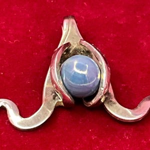 May include: A silver-toned metal pendant featuring a central, smooth, round, light blue stone held within a three-pronged setting. The metal frame extends outward into two symmetrical, curved, wave-like arms that taper to points at the bottom ends.