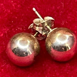 May include: A pair of polished silver-tone ball stud earrings. Each earring features a smooth, spherical design with a reflective metallic surface. The earrings include standard post backs and butterfly-style friction clutches for secure attachment.