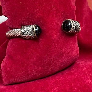 May include: A silver cuff bracelet featuring a twisted cable texture on the band. Each end of the open cuff is capped with a decorative silver filigree setting that holds a smooth, round, polished black stone cabochon at the center.