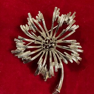 May include: A silver-toned metal brooch shaped like a stylized flower with a textured, radiating petal design. The center features a cluster of small, raised circular details, and a single curved stem extends from the bottom of the floral head.