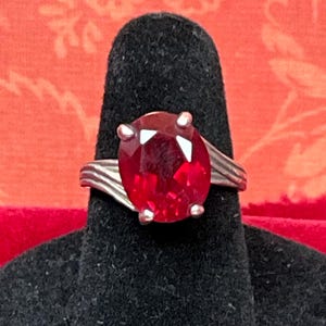 May include: A silver-toned ring featuring a large, oval-cut, deep red gemstone held in place by four prongs. The band of the ring displays a textured, ribbed design that tapers as it extends from the central setting to the sides of the finger.