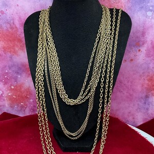 May include: A multi-strand gold-tone necklace featuring several layers of interconnected metal chains. The necklace consists of various chain link styles, including textured and smooth oval links, all hanging in cascading loops of different lengths.