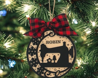 May include: A wooden Christmas ornament featuring a black sewing machine silhouette and the name "ROBIN". It has a red and black plaid bow and is suspended by twine. The ornament is decorated with snowflakes, stars, and trees, and is hanging on a Christmas tree.