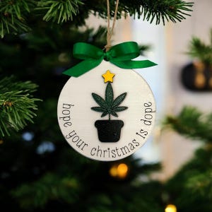 May include: A circular Christmas tree decoration with a cannabis plant design. The white ornament has the text "hope your christmas is dope", a green ribbon, and a yellow star. The plant is in a black pot.