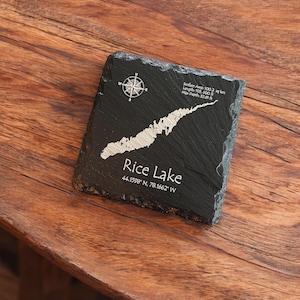 Peut inclure: Un dessous de verre en ardoise noire avec une carte blanche du lac Rice, des coordonnées et une rose des vents. Le dessous de verre porte l'inscription "Rice Lake" et les coordonnées 44.1598° N, 78.1662° W. Surface 100,2 km², longueur 32 187,8 m, et profondeur max 10 m.