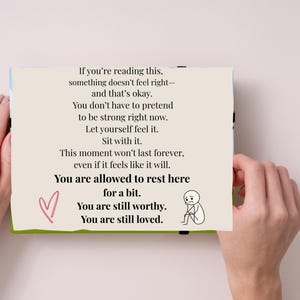 May include: A card with text that reads, "If you're reading this, something doesn't feel right - and that's okay." The card also has a pink heart and a stick figure. The card is held by two hands.